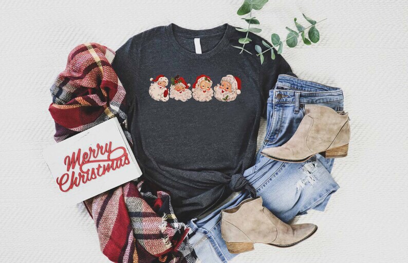 May include: A dark gray t-shirt with a vintage-style print of four Santa Claus faces. The shirt is tied in a knot and is laying on a white surface with a plaid blanket, a pair of jeans, and two brown ankle boots.