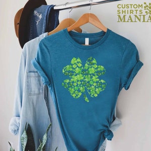 May include: A teal blue t-shirt with a four-leaf clover design made of smaller green clovers. The text "CUSTOM SHIRTS MANIA" is in the top right corner of the image.
