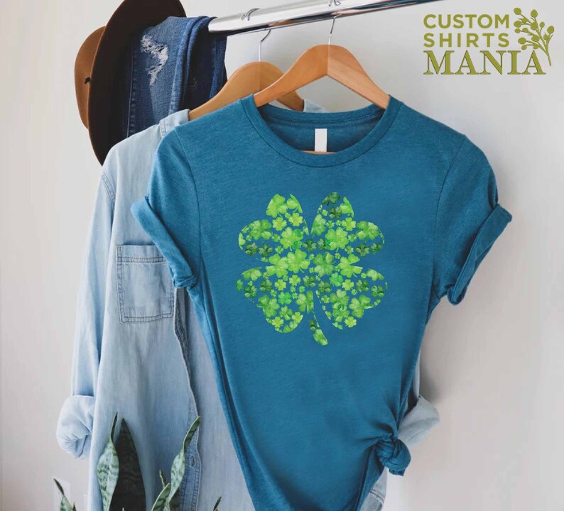 May include: A teal blue t-shirt with a four-leaf clover design made of smaller green clovers. The text "CUSTOM SHIRTS MANIA" is in the top right corner of the image.