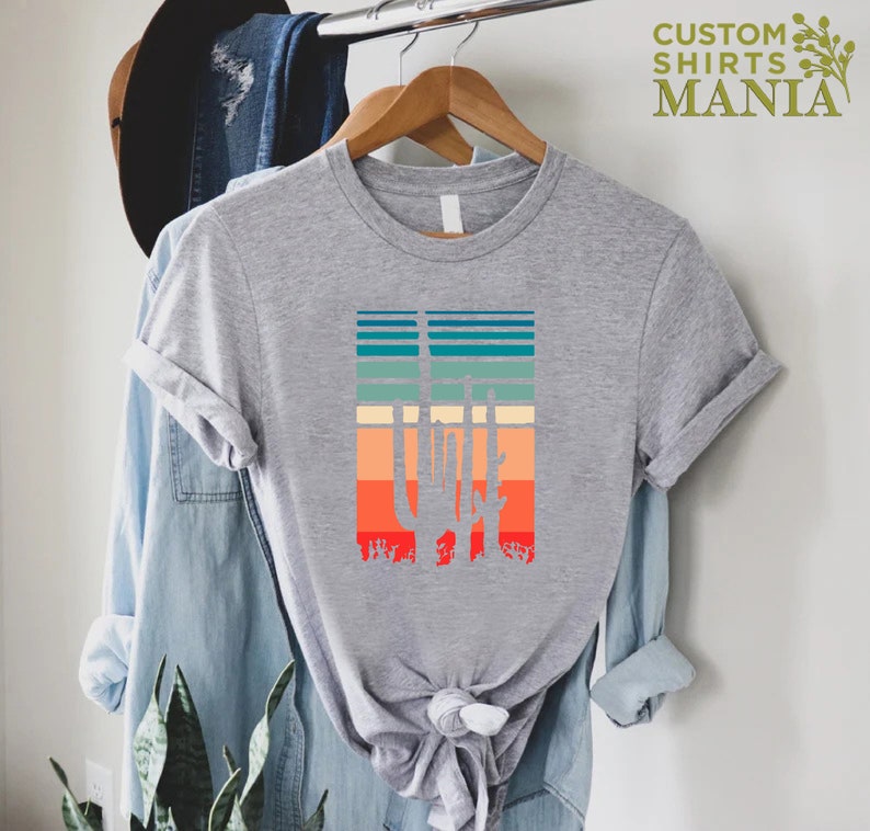 May include: A gray t-shirt with a retro-style graphic of a cactus silhouette against a colorful striped background. The text "CUSTOM SHIRTS MANIA" is visible in the top right corner of the image.