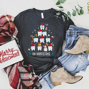 以下が含まれることがあります： 歯でできたクリスマスツリーのデザインが施されたダークグレーのTシャツ。ツリーは、キャンディーケーン、オーナメント、雪だるまなどで飾られています。ツリーの下には「Oh Dentistree」という文字が印刷されています。