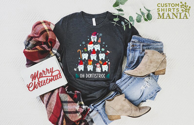 以下が含まれることがあります： 歯でできたクリスマスツリーのデザインが施されたダークグレーのTシャツ。ツリーは、キャンディーケーン、オーナメント、雪だるまなどで飾られています。ツリーの下には「Oh Dentistree」という文字が印刷されています。