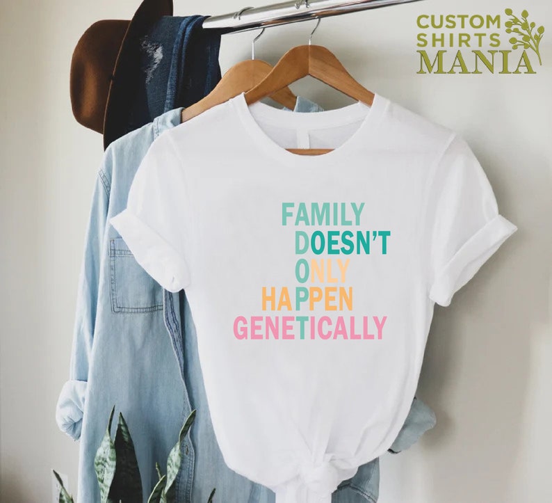Op de afbeelding: Wit T-shirt met een kleurrijke afbeelding met de tekst "Family Doesn't Only Happen Genetically".