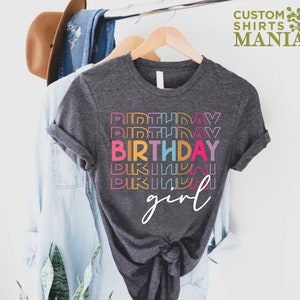 May include: A dark grey t-shirt with the text "BIRTHDAY girl" in rainbow colours. The word "birthday" is repeated multiple times in different colours.