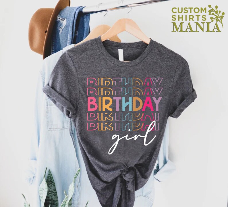 May include: A dark grey t-shirt with the text "BIRTHDAY girl" in rainbow colours. The word "birthday" is repeated multiple times in different colours.