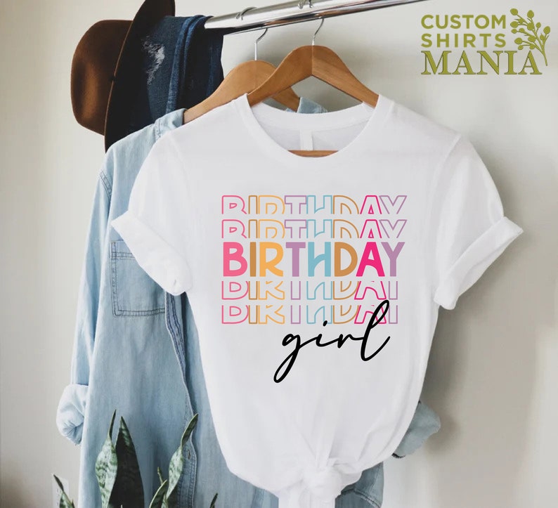 May include: White t-shirt with the text "Birthday girl" in rainbow colours. The text is stacked and the word "Birthday" is repeated multiple times.