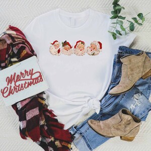 May include: White t-shirt with a graphic of four vintage Santa Claus faces. The shirt is tied in a knot at the bottom. The shirt is laying on a white surface with a plaid scarf, a sign that says "Merry Christmas", and a pair of brown ankle boots and blue jeans.