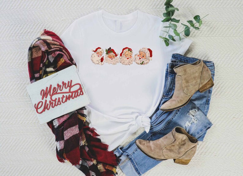 May include: White t-shirt with a graphic of four vintage Santa Claus faces. The shirt is tied in a knot at the bottom. The shirt is laying on a white surface with a plaid scarf, a sign that says "Merry Christmas", and a pair of brown ankle boots and blue jeans.