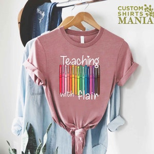 May include: A pink t-shirt with the text "Teaching with Flair" and a graphic of colorful markers.
