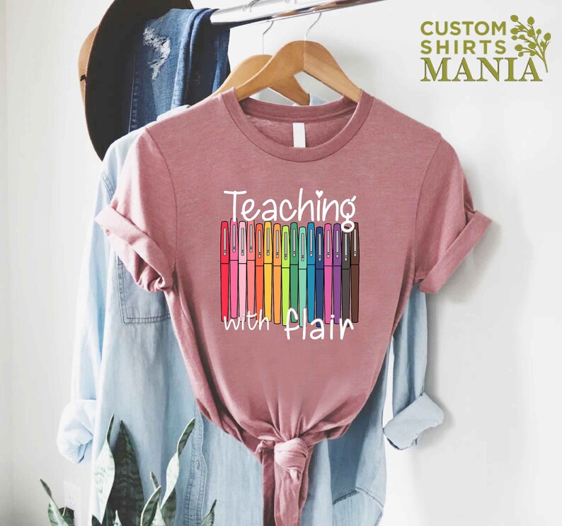 May include: A pink t-shirt with the text "Teaching with Flair" and a graphic of colorful markers.