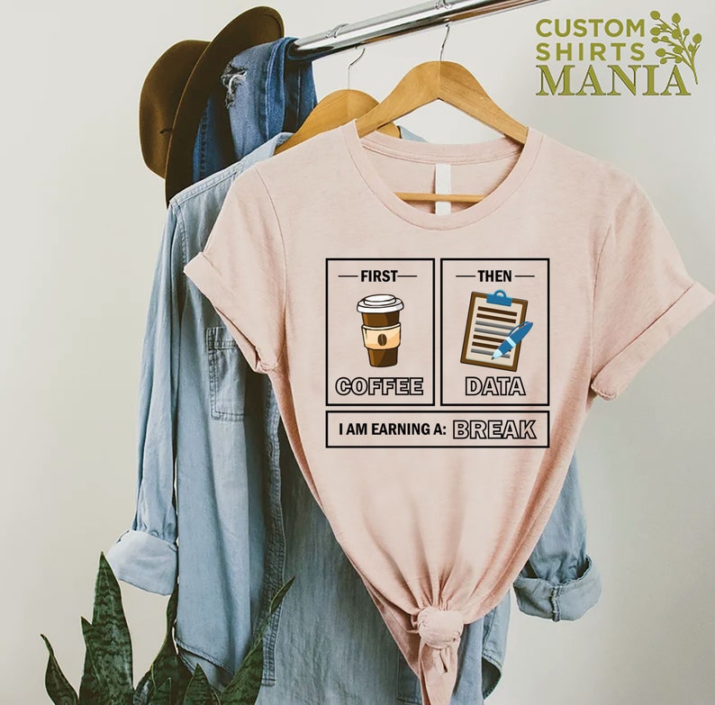 May include: A pink t-shirt with a graphic design that says "First Coffee, Then Data, I am earning a break". The graphic features a cup of coffee and a clipboard with a pen.