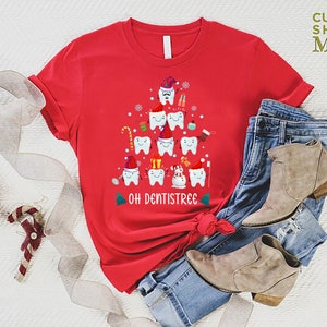 以下が含まれることがあります： 歯でできたクリスマスツリーのデザインが施された赤いTシャツ。ツリーはサンタの帽子、キャンディーケーン、オーナメントで飾られています。ツリーの下には「Oh Dentistree」という文字が印刷されています。