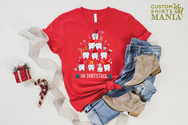 以下が含まれることがあります： 歯でできたクリスマスツリーのデザインが施された赤いTシャツ。ツリーはサンタの帽子、キャンディーケーン、オーナメントで飾られています。ツリーの下には「Oh Dentistree」という文字が印刷されています。