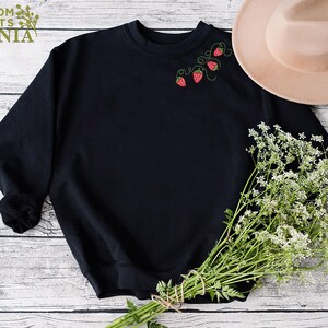 Puede incluir: Sudadera negra con fresas bordadas y vides verdes. El texto "CUSTOM SHIRTS MANIA" es visible en la esquina superior izquierda. Un sombrero beige y un ramo de flores blancas tambi&eacute;n est&aacute;n en la imagen.