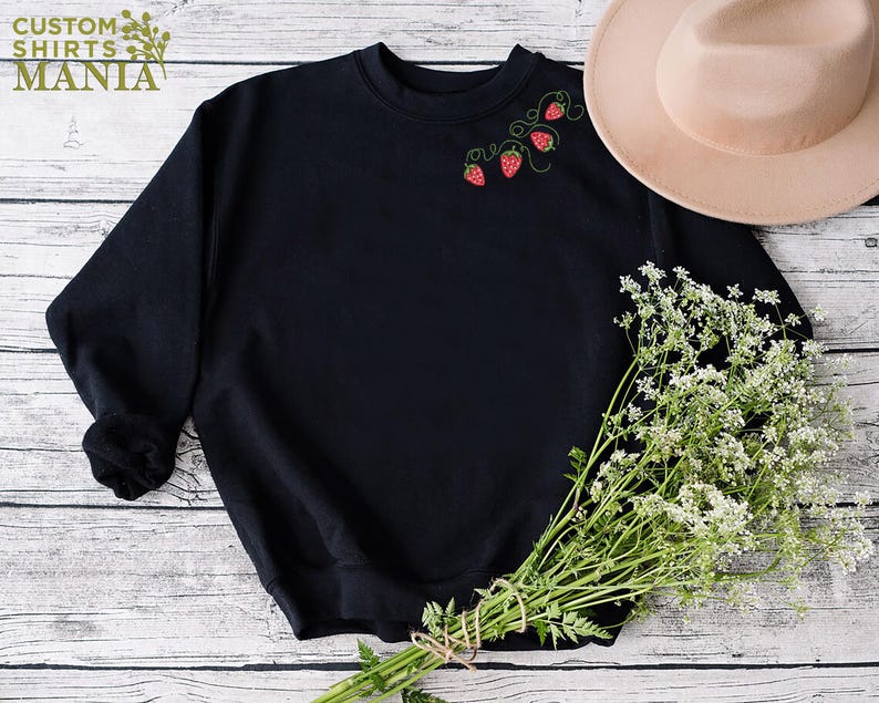Puede incluir: Sudadera negra con fresas bordadas y vides verdes. El texto "CUSTOM SHIRTS MANIA" es visible en la esquina superior izquierda. Un sombrero beige y un ramo de flores blancas tambi&eacute;n est&aacute;n en la imagen.
