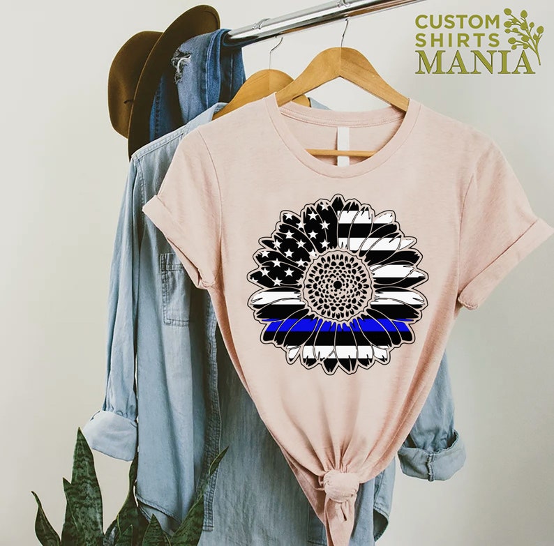 May include: A pink t-shirt featuring a black and white sunflower design. The sunflower has a blue line running through the centre, representing the thin blue line.