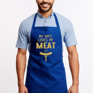 May include: A blue apron with a white pocket and the text "My wife loves my meat" in gold lettering. A gold cartoon hot dog with a fork is below the text.