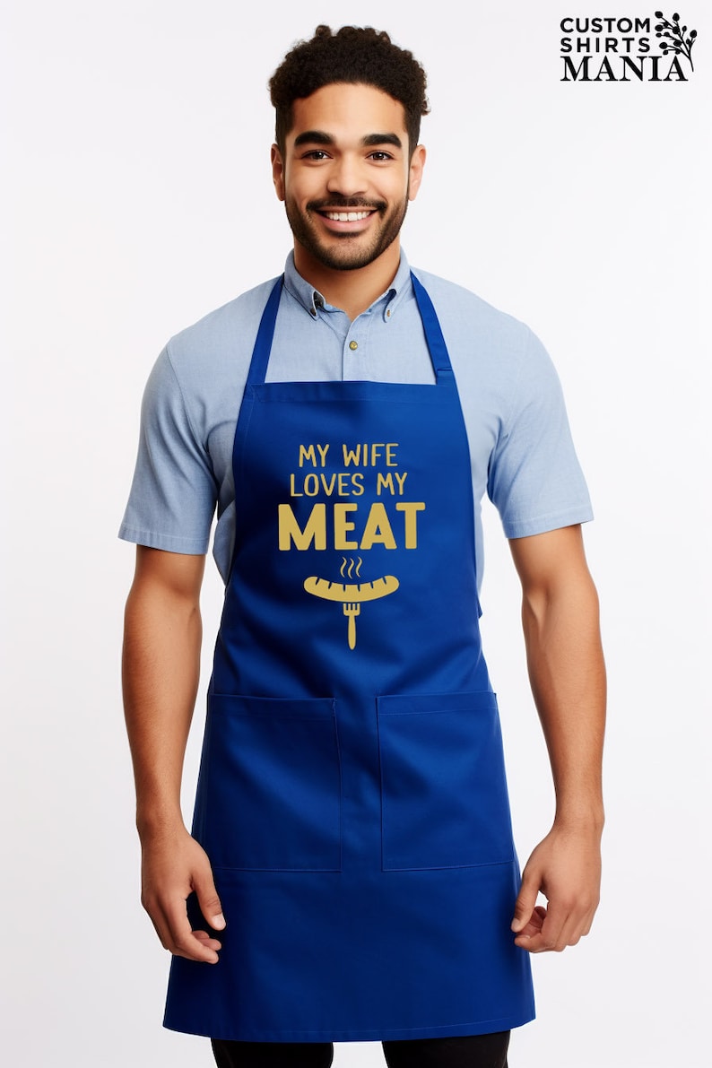 May include: A blue apron with a white pocket and the text "My wife loves my meat" in gold lettering. A gold cartoon hot dog with a fork is below the text.