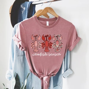 May include: A mauve-colored t-shirt featuring a graphic of three crawfish. The crawfish are in red and pink with decorative patterns. The text "crawfish season" is printed in white below the crawfish.