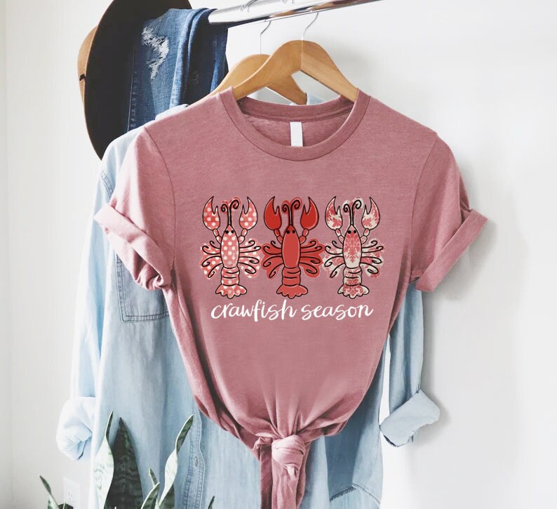 May include: A mauve-colored t-shirt featuring a graphic of three crawfish. The crawfish are in red and pink with decorative patterns. The text "crawfish season" is printed in white below the crawfish.