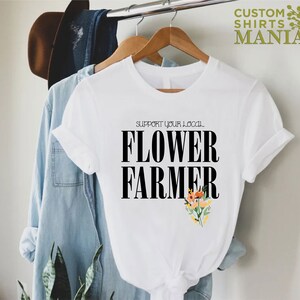 May include: White t-shirt with black text that reads "Support your local Flower Farmer". The text is in a bold font. There are small colorful flowers in the bottom right corner of the shirt.
