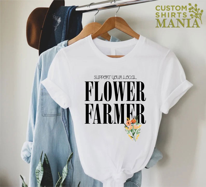 May include: White t-shirt with black text that reads "Support your local Flower Farmer". The text is in a bold font. There are small colorful flowers in the bottom right corner of the shirt.