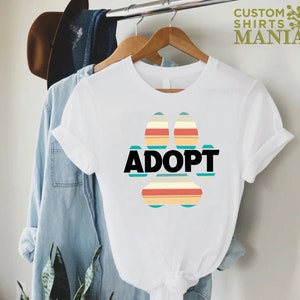 May include: White t-shirt with a colorful paw print design and the word "ADOPT" in black text.