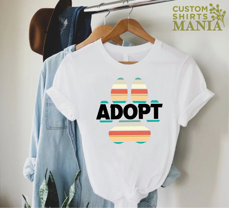 May include: White t-shirt with a colorful paw print design and the word "ADOPT" in black text.