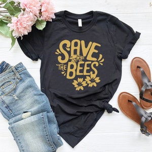May include: Black t-shirt with the text "SAVE THE BEES" in gold lettering, featuring a bee graphic and two flower graphics. The shirt is paired with blue jeans and brown sandals. Pink flowers are in the background.