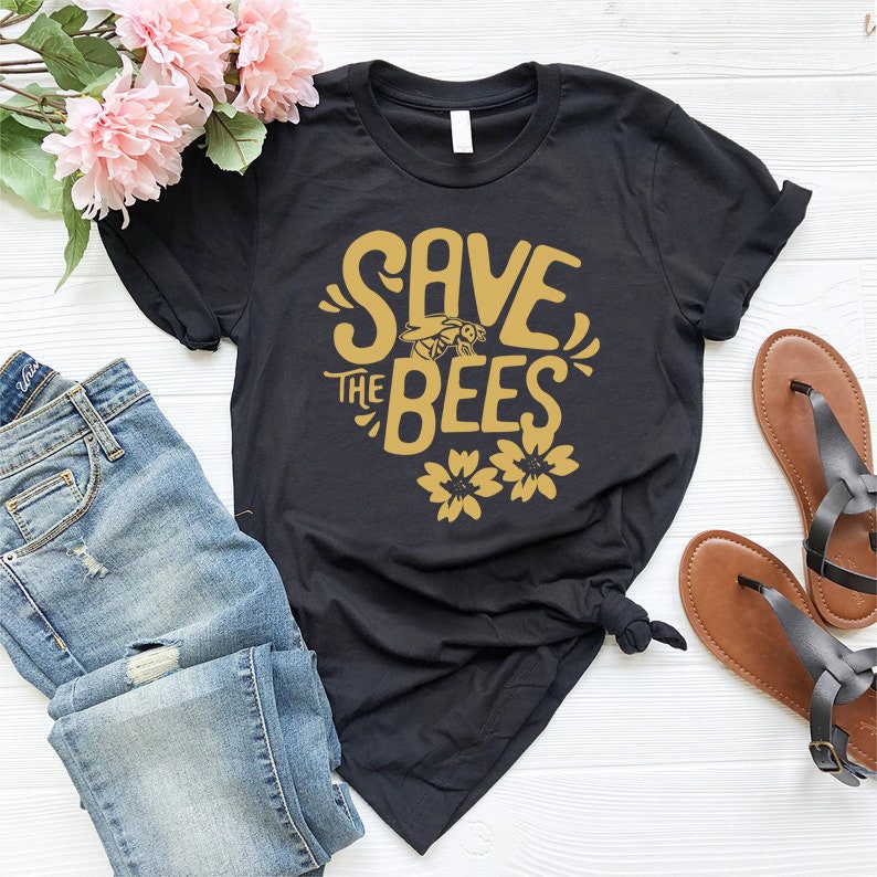 May include: Black t-shirt with the text "SAVE THE BEES" in gold lettering, featuring a bee graphic and two flower graphics. The shirt is paired with blue jeans and brown sandals. Pink flowers are in the background.