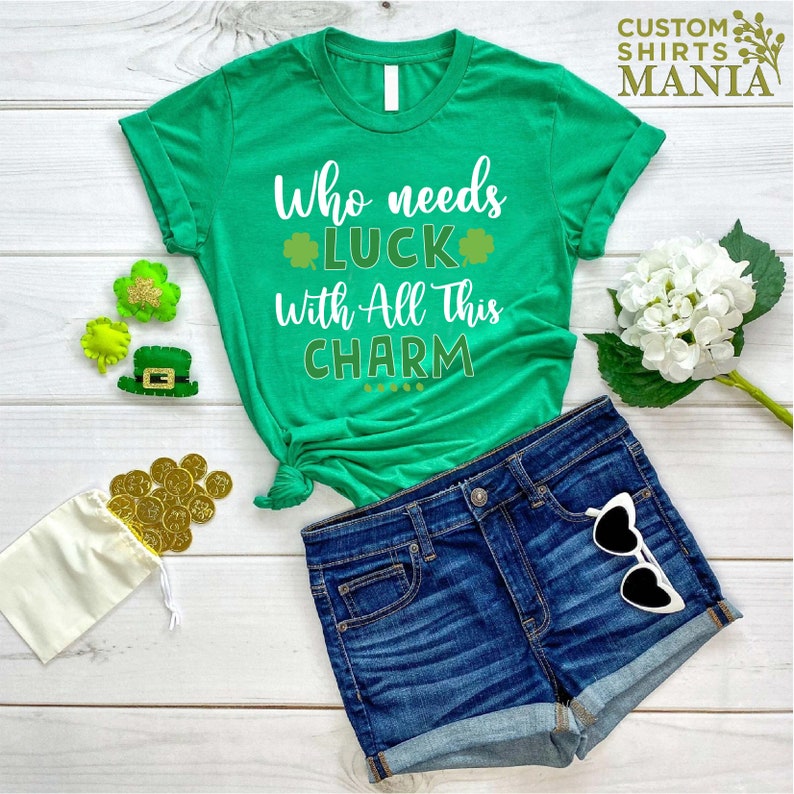 May include: Green t-shirt with white text that reads "Who needs luck with all this charm". The shirt is on a white wooden surface with a pair of blue denim shorts, gold coins, and green shamrock decorations.