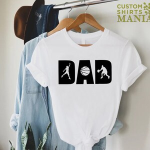 Op de afbeelding: Wit T-shirt met een zwart grafisch ontwerp met de tekst "DAD" met een basketbal en twee basketballers.