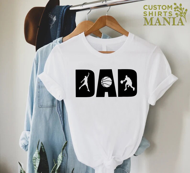 Op de afbeelding: Wit T-shirt met een zwart grafisch ontwerp met de tekst "DAD" met een basketbal en twee basketballers.