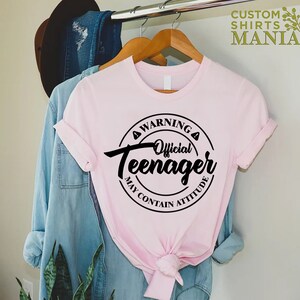 May include: Pink t-shirt with a black circle graphic that says "Warning Official Teenager May Contain Attitude".