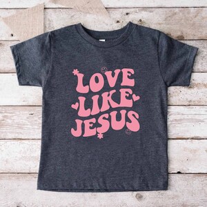 Może przedstawiać: Szary t-shirt z r&oacute;żowym nadrukiem, na kt&oacute;rym widnieje napis "LOVE LIKE JESUS" w retro czcionce. Nadruk zawiera r&oacute;wnież małe r&oacute;żowe serduszka i kwiaty.