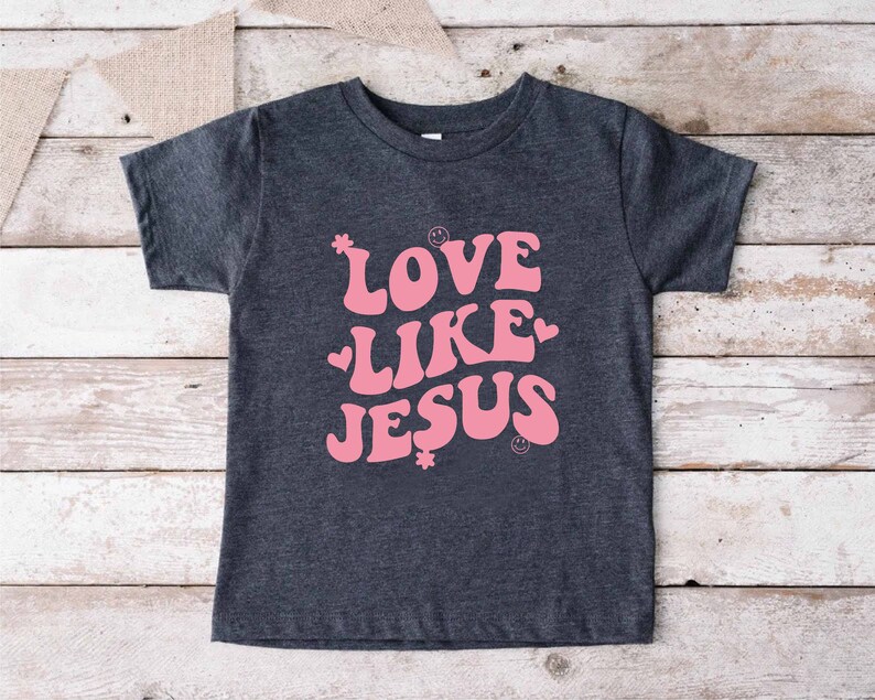 Może przedstawiać: Szary t-shirt z r&oacute;żowym nadrukiem, na kt&oacute;rym widnieje napis "LOVE LIKE JESUS" w retro czcionce. Nadruk zawiera r&oacute;wnież małe r&oacute;żowe serduszka i kwiaty.