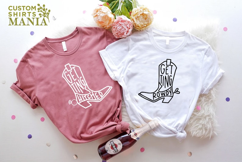 May include: Two t-shirts with a cowboy boot design. One shirt is pink with white text that says "Getting Hitched". The other shirt is white with black text that says "Getting Rowdy".