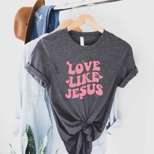 Może przedstawiać: Szary t-shirt z r&oacute;żowym nadrukiem z napisem "Love Like Jesus" w retro stylu.