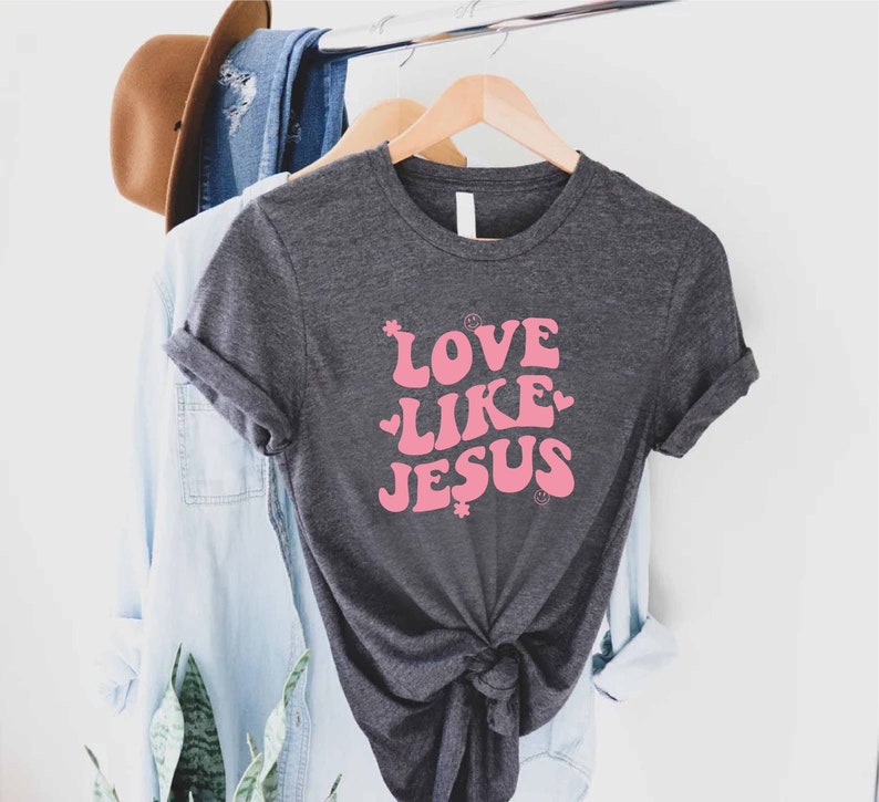 Może przedstawiać: Szary t-shirt z r&oacute;żowym nadrukiem z napisem "Love Like Jesus" w retro stylu.