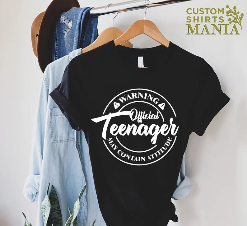 May include: Black t-shirt with a white circle graphic that says "Warning Official Teenager May Contain Attitude".