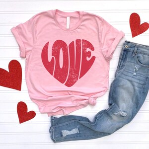 May include: Pink t-shirt with a red heart-shaped graphic that says "LOVE" in a retro font. The shirt is paired with blue jeans.