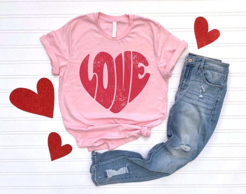 May include: Pink t-shirt with a red heart-shaped graphic that says "LOVE" in a retro font. The shirt is paired with blue jeans.
