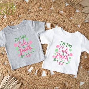 May include: Two toddler-sized t-shirts with the text "I'm Too Cute To Pinch" in pink and green lettering. The shirts are on a woven straw background.