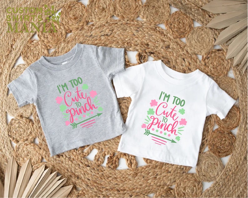 May include: Two toddler-sized t-shirts with the text "I'm Too Cute To Pinch" in pink and green lettering. The shirts are on a woven straw background.