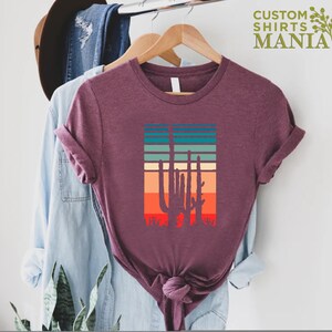 May include: A maroon t-shirt with a vintage-style graphic of three cacti in front of a sunset. The graphic has a retro color palette of orange, red, yellow, green, and blue.