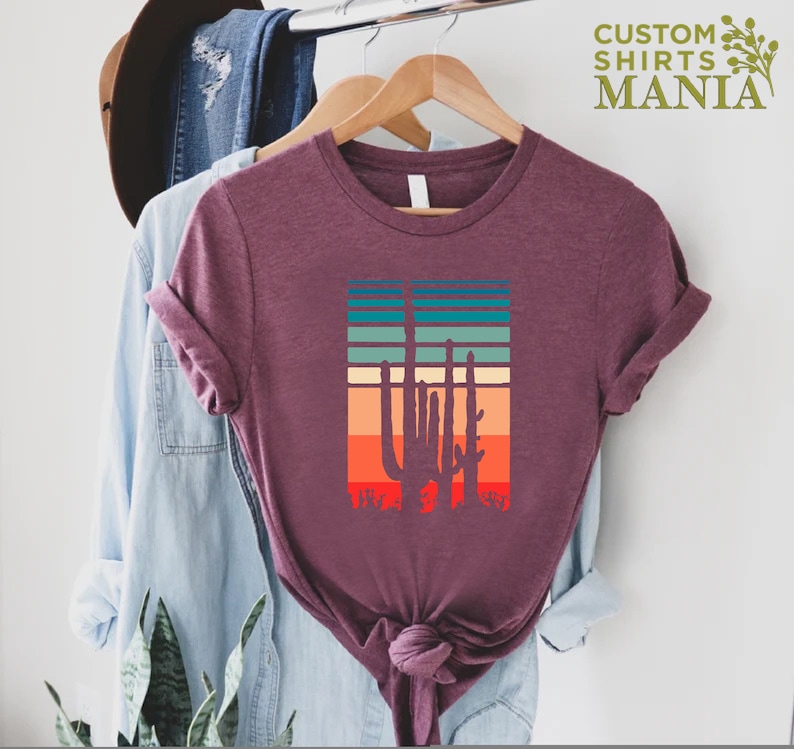 May include: A maroon t-shirt with a vintage-style graphic of three cacti in front of a sunset. The graphic has a retro color palette of orange, red, yellow, green, and blue.