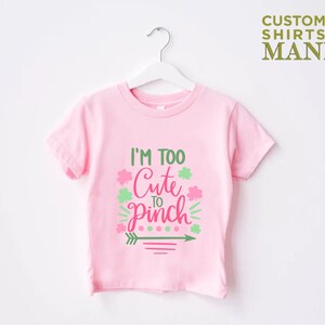 May include: Pink t-shirt with a green shamrock and pink text that reads "I'm Too Cute To Pinch".