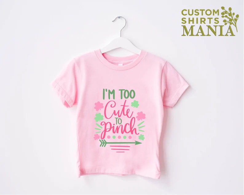 May include: Pink t-shirt with a green shamrock and pink text that reads "I'm Too Cute To Pinch".