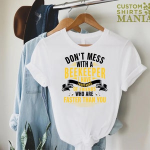May include: White t-shirt with black text that reads "Don't mess with a beekeeper I have thousands of friends who are faster than you". The text is surrounded by a yellow banner with bee silhouettes.