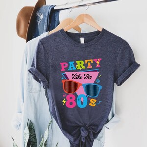 K&ouml;nnte beinhalten: Ein dunkelblaues T-Shirt mit einem grafischen Design, das den Text "Party Like The 80's" in bunten Buchstaben und eine Retro-Sonnenbrille zeigt.